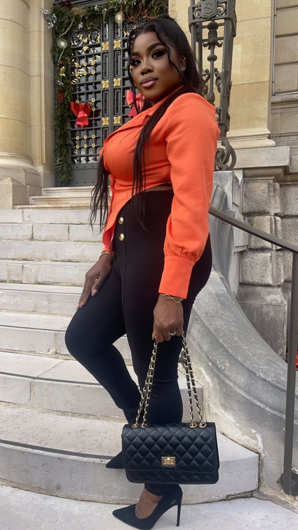 Jinette en blazer orange et pantalon noir, sac matelassé à la main, sur des marches de pierre
